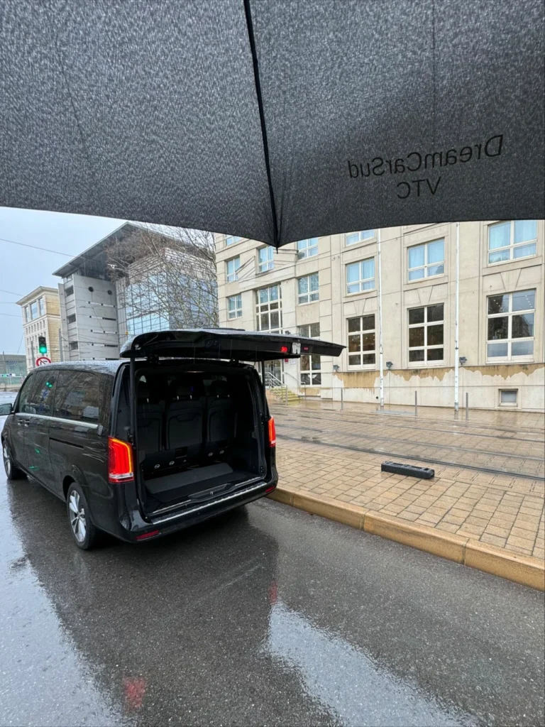 Vue depuis sous un parapluie d'un Mercedes V-Class avec le coffre ouvert, garé dans la rue devant un bâtiment, par un jour pluvieux.