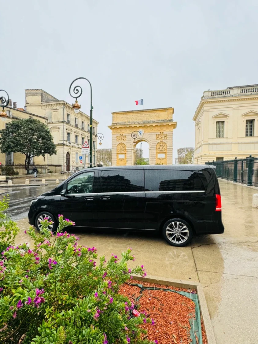 Mercedes V-Class noire stationnée près d'une place ornée de fleurs et d'un monument, dans une ville par un jour de pluie.