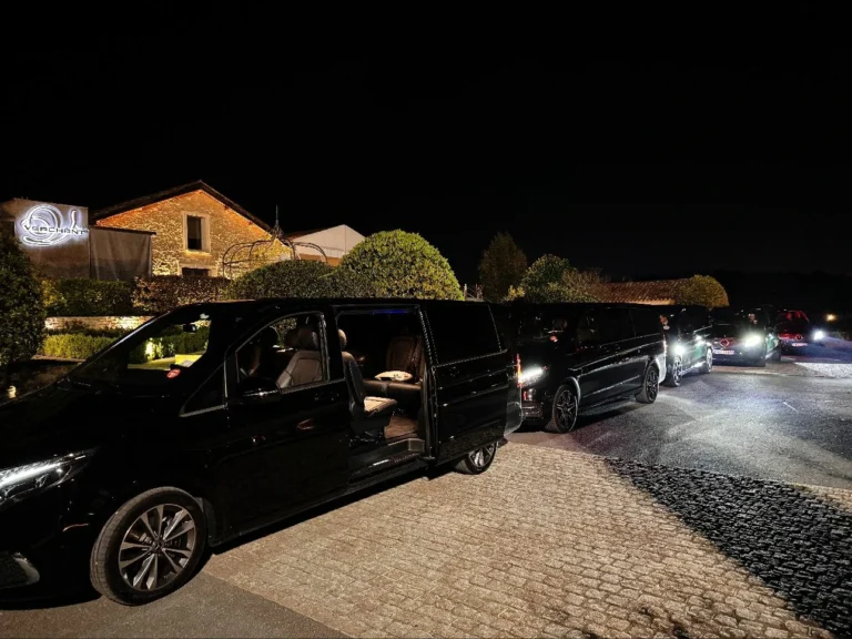 Un Mercedes V-Class noir avec les portes ouvertes, éclairé de nuit, avec d'autres véhicules alignés en arrière-plan devant un bâtiment avec des lumières.